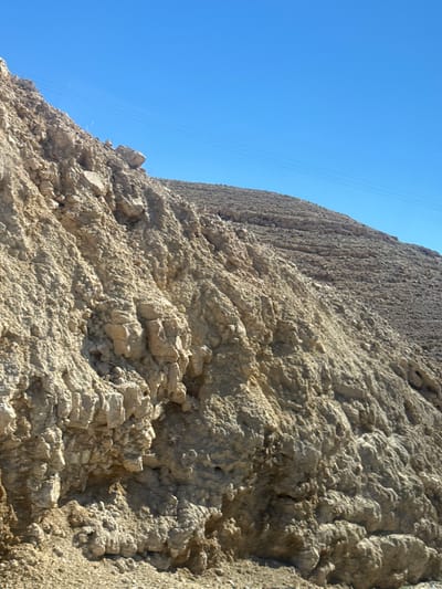 The image shows a rugged, tan-colored cliff face under a clear blue sky. The rock is heavily textured with crevices, small boulders, and erosion patterns. There are thin power lines that run diagonally across the blue sky in the upper left corner of the image. Based on the location, it is likely to be near Arad, Israel. The scene depicts a natural environment with prominent geological features.