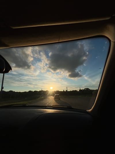 The image depicts a forensic view from inside a moving or stationary passenger vehicle, looking forward through the windshield at sunset in Tovstoluh, Ukraine. The dashboard occupies the lower third of the frame, dark and featureless. The interior roof lining and front sun visors are visible at the top, framing the windshield opening. A rearview mirror is partially visible on the upper left.

The exterior scene is dominated by a low-angle sun, positioned slightly right of the central horizon, emitting intense light that creates significant lens flare and glare on the windshield, highlighting numerous dust particles or water droplets. The sky transitions from a brilliant gold and light blue near the sun to a deeper blue further away, populated by scattered cumulus and cirrus clouds.

A two-lane paved road extends into the distance. Ahead, a dark-silhouetted vehicle, consistent with an SUV or similar, is visible, partially obscured by the sun's glare. The horizon line is characterized by a dense, dark silhouette of trees and vegetation on both sides of the road. Several utility poles are visible along the roadside. No human subjects are directly depicted. The overall lighting is high contrast, with the brightly illuminated exterior and the largely underexposed vehicle interior.