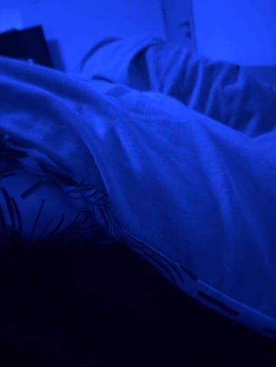The scene depicts an indoor environment in Nadarevo, Bulgaria, illuminated by a pervasive deep blue artificial light source. A human subject is present, lying in a recumbent position, mostly obscured by white bedding that exhibits numerous folds and creases. An underlying fabric, likely a pillow or a secondary sheet, displays a dark linear or abstract pattern visible along the left and lower-left edges of the frame. The background consists of a light-colored wall and indistinct, dark shapes, possibly furniture or electronics, which are blurred due to low light and shallow depth of field. The lower foreground is uniformly dark, indicative of deep shadow. No specific actions, interactions, or identifying features of the subject are discernible beyond their static presence under the covers.