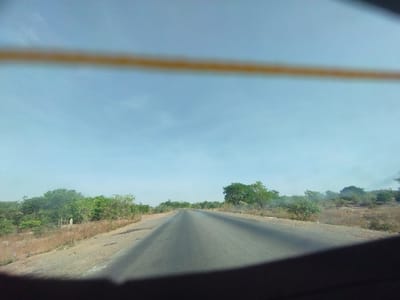 The image is a view from a vehicle's rearview mirror. It shows a paved road with a dividing line in the middle. The road is empty. There are trees and bushes on either side of the road. The sky is clear with a few white clouds. The coordinates provided (latitude: 9.71, longitude: 11.419) place the location in Lakweme, Nigeria. The scene is a typical rural landscape with a paved road running through it. There are no people or objects in the foreground, only the road and vegetation. The image suggests a peaceful and quiet environment.