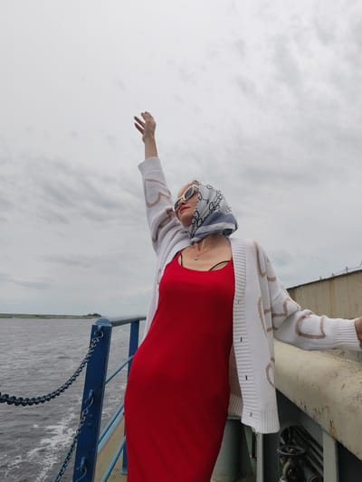 A woman stands on the deck of a boat, her right arm raised towards the sky, her fingers spread. She is wearing a red dress and a white cardigan with a heart pattern, a patterned scarf on her head, and white sunglasses. The scene appears to be a cloudy day, with a body of water on the left and a railing on the right. The woman's pose suggests a sense of freedom or exhilaration. The location is identified as Bereg, Russia.