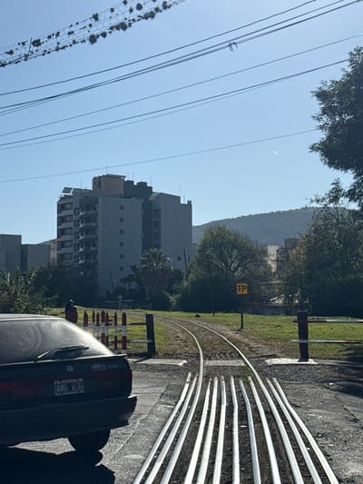 The image shows a street scene in Salta, Argentina. In the foreground, there are railway tracks curving to the right. To the right of the tracks, a dark-colored car is parked, with a license plate visible, reading "AMG 836". Beyond the tracks and car, there is a grassy area with posts and a yellow sign marked "TP". In the background, there are tall buildings and a tree-covered mountain. Power lines run along the left side of the image.