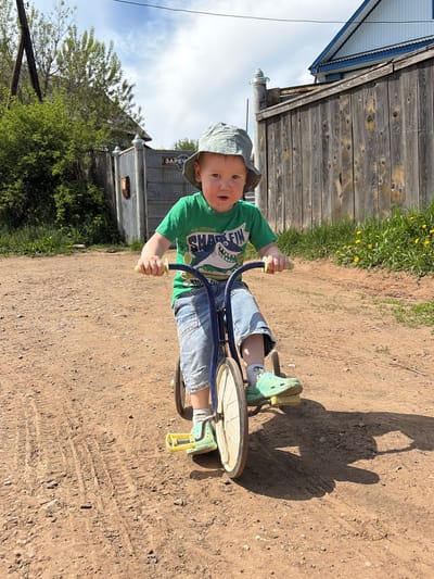 A young child wearing a green t-shirt, blue jeans, and a light-colored hat is riding a tricycle on a dirt surface. The tricycle has blue handlebars and a large front wheel. The child is wearing green Crocs-style shoes with yellow accents. The background includes a wooden fence, green vegetation with yellow flowers, a building with a metal exterior, and a partly cloudy sky. The shadow of a person holding a camera is cast on the dirt surface. The location is possibly near a rural area in Ul'yanovka, Russia, based on the Cyrillic writing "ЗАРЕЧ" on the metal building.