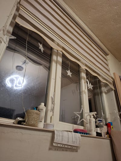 This image depicts an indoor window scene, likely in an apartment in Belozem, Bulgaria. The window is covered by a horizontally striped roller blind with alternating opaque and sheer bands. Dangling from the blind and window frame are several clear, star-shaped LED lights, suggesting a festive or decorative display.
On the windowsill, various items are arranged. To the left, a woven basket holds what appear to be pens or markers. Next to it, a white bottle with a pump dispenser is present, along with a cylindrical container. Further to the right, another white bottle, this one with a spray nozzle, stands prominently. Behind it, a box labeled "FOLIAIN Minoxidil" is visible, along with other small containers and a bright blue spray can. A pink object rests on a blue ribbed mat.
The window itself is dirty, with streaks and smudges obscuring the view outside. However, a bright white, circular neon-like light is reflected on the glass, forming a looped or stylized "S" shape. The wall below the windowsill is textured and appears to be painted a light color, with some water stains or discoloration visible. A folded document with printed text and checkboxes is clipped to the wall below the windowsill to the left, and another document is partially visible on the right. The overall impression is one of a utilitarian space decorated for a special occasion.