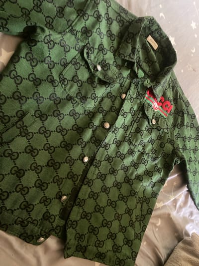 The image displays a green garment, possibly a jacket or shirt, laid flat on light gray bedding adorned with white star and crescent moon patterns. To the right, a soft, lighter-colored fabric, likely a blanket or pillow, is partially visible.
The garment itself is patterned with a repeating black monogram, featuring the interlocking "GG" logo. It has a classic collar with a white label sewn inside that clearly reads "Gucci" in black text. The garment features a button-front closure and two chest pockets, each secured by a silver-toned button. On the left chest pocket (viewer's perspective), a red and green striped ribbon is embroidered, with additional red embroidery on top that appears to be "50" or "SG," accompanied by a small gold or white element within the stripe. The buttons on the garment are round and silver-toned, each featuring an embossed design that is difficult to discern precisely but resembles intertwined letters or a crest. The fabric of the garment appears to be textured.
No individuals are present in the image, and no activity is occurring; the garment is stationary. The setting appears to be indoors, likely a bedroom, suggested by the bedding.