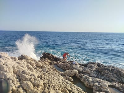 In Karaburun, Turkey, a person wearing a red t-shirt and a straw hat is crouched on the rocky shore of the Aegean Sea. The sea is a deep blue, with waves crashing against the rocks, creating a spray of white foam. The rocks are rough and varied in color, ranging from light tan to dark grey. In the background, the horizon is a clear, light blue sky. A small boat is visible in the distance on the sea. The overall scene depicts a natural coastal environment with a human presence.