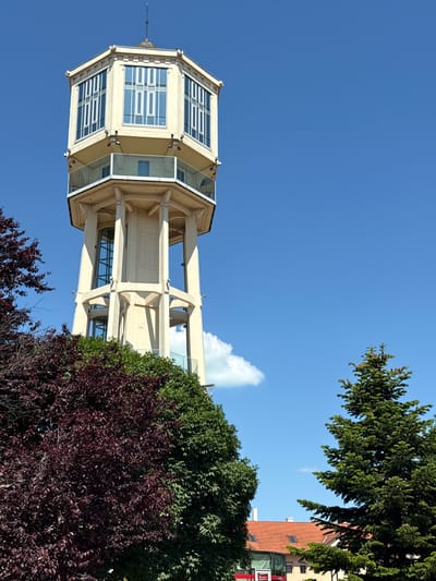 The image depicts the Siófok Water Tower, a prominent landmark in Siófok, Hungary. The tower is a tall, light-colored structure with a distinctive, multi-windowed top section. It is viewed from a low angle, emphasizing its height against a clear blue sky. A few trees and bushes are visible in the foreground, partly obscuring the tower's lower portion. Part of a building can be seen to the right.