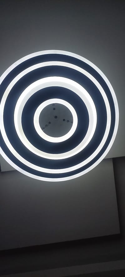 This is a close-up, low-angle shot of a modern, circular ceiling light fixture with concentric rings. The fixture has alternating dark blue and bright white rings, with the white rings appearing to be illuminated. The center of the fixture is visible and shows a light gray surface with several small, dark circular or dot-like details, possibly screw heads or mounting points. The background is a plain, gray wall that is partially in shadow, suggesting an indoor setting. The lighting is primarily from the fixture itself, creating bright highlights on its surface and casting soft shadows on the wall. The overall mood is clean and contemporary. There is no text visible in the image. The location context is Laminga, Nigeria.