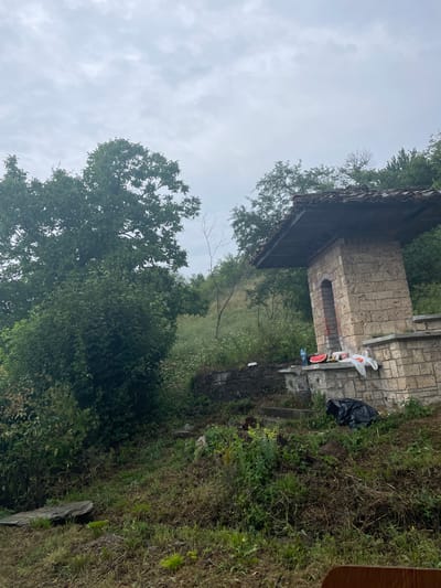 In the village of Ivanovtsi, Bulgaria, a stone structure with a tiled roof, resembling a small shelter or monument, stands amidst lush greenery under a cloudy sky. To the right of the structure, on a stone ledge, are remnants of a meal: a sliced watermelon, a bottle of water, and some plastic bags. A black trash bag rests on the ground at the base of the structure, suggesting recent activity or a picnic. The surrounding landscape is densely vegetated with trees and wild grass, with a stone pathway leading up to the structure. The overall scene conveys a sense of a rustic outdoor location, possibly a rest stop or a historical marker.