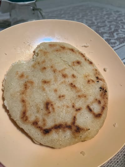 A close-up shot shows a single, cooked arepa resting on a light pink plate. The arepa is round and slightly flattened, with a golden-brown color and grill marks indicating it was cooked on a griddle. There are some darker, charred spots on its surface. The plate is slightly tilted, and a patterned tablecloth is visible in the background. The scene is indoors, likely in a home setting. The presence of an arepa suggests a location in Venezuela, potentially the city of Villa de Cura, where arepas are a staple food. The lighting is soft and natural.