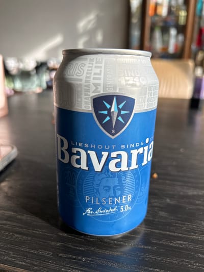 A blue and silver beer can sits on a dark wooden table. The can is branded "Bavaria Pilsener" and has the slogan "Lieshout Sinds 1719". The background is blurred, showing a domestic interior with a cabinet filled with various items and a chair. The lighting suggests a natural light source coming from the left, casting shadows on the table and the can. There is no indication of the location being in the city of Boxmeer, Netherlands.