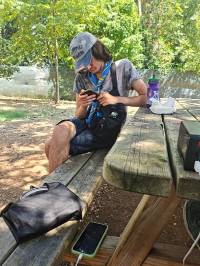 A young male with long, dark hair and a blue and yellow scarf around his neck is sitting on a wooden picnic table bench, looking at his smartphone. He is wearing a grey t-shirt and dark shorts, and a grey baseball cap. A black crossbody bag is slung over his shoulder. On the table, there is a purple water bottle, a white packet, and a black portable charger connected to a green-cased smartphone. Another black bag is on the bench next to him. The background shows greenery and trees, suggesting an outdoor setting. The scene appears to be in a park or recreational area, possibly within the city of L'Aquila, Italy, given the context provided by the user.