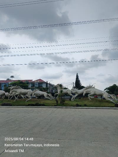 The image presents an outdoor scene from Pusakarakyat, Kecamatan Tarumajaya, Indonesia, captured at 14:48 on August 4, 2025. The central feature is a large, white or light-colored sculpture depicting a group of approximately eight horses in dynamic running or leaping poses, positioned on a green, landscaped mound. In the foreground, a textured concrete road surface occupies the lower half of the frame. The background reveals multi-story buildings with red roofs, various trees including a prominent tall conifer, and a faint red vehicle in the distance. The sky is overcast with grey clouds, and multiple horizontal overhead utility lines are visible across the upper portion of the scene. No individuals are present.