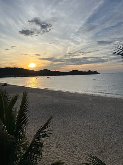 The image captures a serene beach scene at sunset in Lệ Uyên, Vietnam. The sun, a brilliant orb, is partially obscured by rolling hills in the distance, casting a warm, golden glow across the water and the sky. The sky is a canvas of soft oranges, yellows, and blues, with wispy clouds stretching across it. The calm sea reflects the sunlight, creating a shimmering path from the horizon to the shore. A wide, sandy beach stretches across the foreground, its surface textured with the gentle ripples left by the receding tide. In the distance, several small boats are anchored, their silhouettes adding a touch of human presence to the natural landscape. The foreground is framed by the dark, silhouetted fronds of a palm tree, adding depth and a tropical feel to the composition. A red vehicle is parked on the sand near the water's edge, its presence a subtle reminder of the nearby civilization.