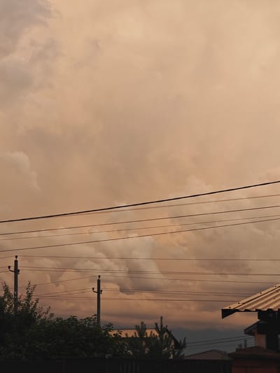 The image depicts a sky filled with dramatic, overcast clouds in a peachy-orange hue, suggesting either sunrise or sunset. Several power lines are stretched horizontally across the frame, supported by two utility poles visible in the middle ground. Below the power lines, the tops of trees and some houses are visible, with the metallic roof of a building on the right. The overall setting appears to be a residential area. The scene does not show any people or indicate any specific actions or interactions. The location is identified as Koledino, Russia.