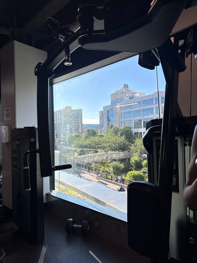 This is a medium shot, taken indoors at a gym, looking out a large window at an urban park setting. The foreground is dominated by gym equipment, with a portion of a weight machine in black metal and padding visible on the right side, and another machine on the left. A single dumbbell rests on the dark gym floor in the lower center.

The window provides a view of a pedestrian walkway with several people walking, a bridge crossing over a vegetated area with trees and rocks, and beyond that, buildings of varying heights, some with visible water towers. The sky is a clear blue. The lighting suggests it is daytime, with sunlight streaming in and casting shadows on the gym floor. The overall impression is one of a well-equipped gym with a scenic urban view.