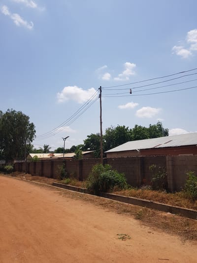 The image depicts an outdoor scene in Palade, Nigeria, under a bright, partly cloudy sky, suggesting it is daytime. The composition is dominated by a dusty, reddish-brown path in the foreground, running along the right side of the frame.

To the left of the path is a low concrete wall, topped with dry, yellowish grass and some green vegetation. Behind the wall, a building with a corrugated metal roof and concrete block walls is visible. Trees with dense green foliage grow behind the building and along the wall, providing shade and a natural element to the scene.

A prominent wooden utility pole stands in the middle ground, supporting several black electrical wires that stretch across the frame. One of these wires has a small, rectangular object attached to it, possibly a bird or a marker. Further down the path, a modern street light with a solar panel is visible, mounted on a pole.

The overall impression is of a somewhat rural or semi-urban environment, typical of many parts of Nigeria. The lighting suggests a sunny day, possibly late morning or early afternoon. No people are visible in the frame. There is a small sign near the bottom left, but the text is too blurry to read.