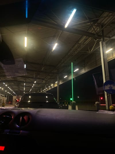 The image depicts a nighttime scene at a border crossing, likely Terespol, Poland. The perspective is from inside a vehicle, looking forward through the windshield. The subjects are primarily the infrastructure of the border crossing and the vehicle's interior.
The scene is characterized by a long, covered structure with a metal framework supporting a corrugated roof. Numerous fluorescent lights illuminate the area, creating a bright, artificial environment. A green light bar, indicating permission to proceed, spans across the lane. To the right, traffic lights with red and green signals are visible, along with automated barriers or toll gates. A dark-colored car is ahead in the lane. The interior of the vehicle shows part of the dashboard, including air vents.
The time of day is clearly night, as indicated by the darkness outside the illuminated area and the artificial lighting. The weather is not discernible from the image. The location is suggested by the context of a border crossing with clear traffic control signals and infrastructure. There are no visible people.
Notable details include the stark contrast between the bright lights and the dark night sky. The arrangement of the lights and the structure suggest a functional, utilitarian space designed for efficient vehicle processing. The image conveys a sense of transition and movement, as vehicles approach and pass through the crossing.
There is a circular sign with a blue background and a white lightning bolt symbol on it, positioned above the traffic signals. Below the green light bar, there is a sign with a red circle and a horizontal white bar, indicating a prohibition or stop. Some illuminated displays with numbers are visible on the right side of the frame, possibly indicating queue numbers or payment information.