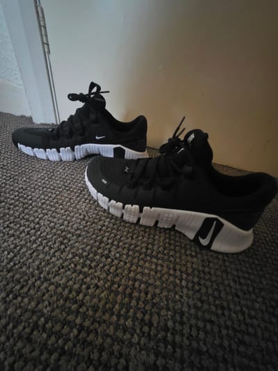 The image displays two black and white Nike athletic shoes positioned on a dark gray, textured carpeted floor. Both shoes feature black uppers with black laces tied, and segmented white soles with a distinctive pattern. A white Nike "Swoosh" logo is visible on the lateral side of the heel of the shoe in the foreground and on the lateral midfoot of the shoe in the background. The word "NIKE" is printed on the tongue of the shoe in the foreground. The shoe in the foreground is angled slightly to the right, with its toe pointing towards the right edge of the frame. The shoe in the background is positioned further left and slightly behind the foreground shoe, oriented in a similar direction. The immediate background consists of a plain, light-colored wall, possibly cream or off-white, which shows minor surface imperfections. To the far left, a white door frame or jamb is partially visible, with a metal door hinge exposed on its vertical edge. The scene is illuminated by ambient indoor lighting, casting soft shadows.