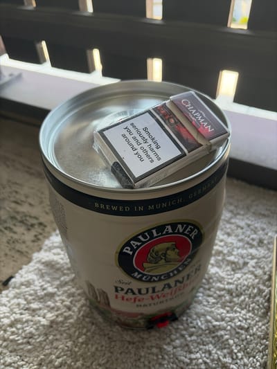 This is a close-up, angled shot of a pack of Chapman cigarettes placed inside the opening of a large can, likely a beer can due to the visible text "PAULANER" and "BREWED IN MUNICH, GERMANY". The can has a pale exterior with a dark band around the rim, and a circular logo featuring a bearded man's profile. The cigarettes are in a dark red and black pack with "CHAPMAN" prominently displayed in white lettering. A health warning in English is visible on the cigarette pack, stating "Smoking seriously harms you and others around you". The background consists of a textured, light-colored surface, possibly a carpet or rug, and some out-of-focus dark shelving with bright horizontal elements, suggesting an indoor setting. The lighting appears to be ambient, creating soft shadows. The scene is static, with no activity taking place beyond the arrangement of the objects. The overall impression is one of a casual, possibly domestic, setting.