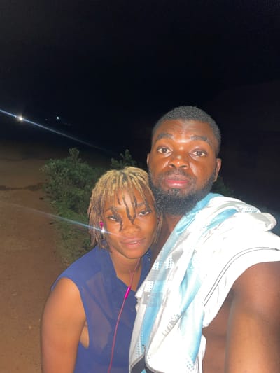 A man and woman are in a selfie photograph taken outdoors at night in Ogbe, Nigeria. The man has a beard, and he is wearing a white garment. The woman has blond dreadlocks and is wearing a blue top. Pink earphones are visible around her neck. Trees are visible in the background.