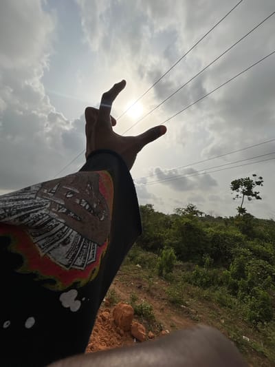 The image depicts a low-angle outdoor scene in Ilishan, Nigeria, viewed upwards towards the sky and horizon. In the immediate foreground, a dark-skinned human left arm, covered by a black long-sleeved garment, extends towards the upper center. The hand is positioned with fingers slightly curved, appearing to "hold" or "pinch" the bright, partially obscured sun between the thumb and forefinger. The garment's sleeve features a graphic design comprising a skull-like image made of what appears to be fanned dollar bills, accented with red and white elements, possibly resembling flames, on a black background.
The sky occupies the upper two-thirds of the frame, characterized by significant white and grey cloud cover. The sun, a bright circular disc, is centrally positioned, emitting a lens flare. Three dark, parallel power lines traverse the sky horizontally in the middle ground, extending from the left past the hand towards the right.
The background, predominantly in the lower right quadrant, reveals a lush, green, hilly landscape. This terrain is densely covered with various types of foliage and bushes. A distinct, sparsely leafed tree stands vertically on the far right against the sky. Patches of reddish-brown earth and scattered rocks are visible in the foreground along the bottom and lower right, suggesting bare ground or a roadside.