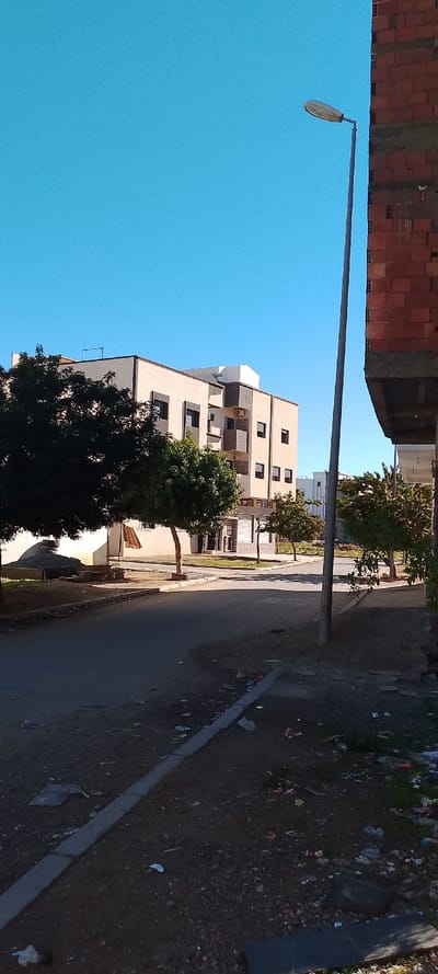 The scene depicts a clear, sunny daytime view of a street in Aïn El Aouda, Morocco. The sky is a uniform, bright blue.
In the immediate foreground on the right, a portion of an unfinished building constructed of red bricks and grey concrete elements is visible, extending vertically out of frame. A grey metallic streetlamp post with an oval-shaped luminaire stands slightly to the left of this building, dominating the mid-right.
The center of the image features a dark asphalt road, slightly curving leftward, with a visible curb lining its right edge. Both sides of the road are characterized by dirt and gravel surfaces heavily strewn with various forms of refuse, including plastic, paper, and general debris.
On the left side of the road, several trees with dark green canopies partially obscure a multi-story building with a light beige facade and darker window frames and balconies. A white structure is visible on the building's roof. Near the base of the trees on the left, a pile of grey aggregate material is present, along with a brown corrugated sheet leaning against a structure. Further down the street, additional buildings and trees are visible in the background. No individuals or visible human activity are present in the image.