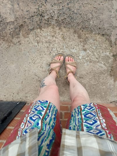 This image shows a close-up, high-angle view of a person's legs and feet, standing on a concrete surface. The person is wearing tan-colored sandals with fabric straps. They are also wearing shorts with a patterned fabric that includes geometric designs in blue, white, and green, bordered by red and brown. The background is a rough, textured concrete surface, possibly an outdoor patio or walkway, with some small stones and darker patches suggesting moisture or dirt. The lighting appears to be natural, indicating daytime. A faint tattoo is visible on the person's left shin. The overall impression is casual, as if someone is posing for a photograph outdoors.