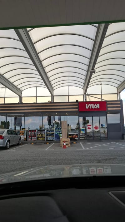 The image captures the exterior of a VIVA convenience store and gas station located in Shtarkovo, Bulgaria. The building features a large, arched translucent canopy providing shelter over a parking area. The store front is characterized by large glass windows and doors. Prominently displayed on the facade is the red VIVA logo. Inside the store, visible through the windows, are shelves stocked with various products, including beverages and other convenience items. To the left of the store, a silver sedan is parked. In front of the store, a wooden pallet with an orange and white traffic cone is situated on the asphalt. The overall environment suggests a typical day at a roadside retail establishment.