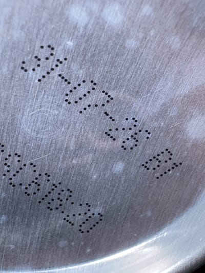 The image depicts a close-up, angled view of a brushed metallic surface, consistent with the lid or base of an aluminum container, likely a beverage can. The surface is grey with a visible linear brushed texture and numerous fine scratches. Several irregularly shaped, lighter grey or white smudges and specks are scattered across the surface. A faint, curved concentric ridge is partially visible at the bottom right, indicating the edge of a circular object.

Three distinct lines of dot-matrix printed text are present:
1.  In the upper-center, angled slightly, the text reads "L19137".
2.  Positioned below and to the right of the first line, the text reads "07:34".
3.  Located below and to the left of the first line, the text reads "2618.20".

All text is rendered in black dots. No people, actions, or interactions are depicted. The environment is limited to the immediate surface of the object. The image provides no information regarding a location in Peschiera Borromeo, Italy, or any other specific geographical context.