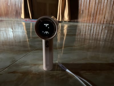 A light pink handheld fan with a digital display is positioned on a marble-patterned tiled floor. The fan has a circular head with a dark screen displaying "AIR VOLUME" with an icon of air flow, and "POWER" indicated by three illuminated white dots, suggesting a medium power setting. A white lanyard is attached to the bottom of the fan. In the background, the fan is positioned in front of tall, vertical wooden blinds and a floor reflecting the blinds and the fan. The scene appears to be indoors, potentially in Turumame, Indonesia.