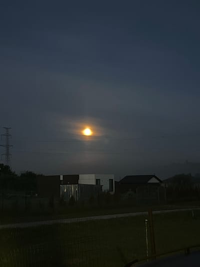 A wide shot captures a twilight scene in Astomi, Latvia, dominated by a dimly lit sky with a bright, hazy celestial body, possibly the moon, at the center, partially obscured by clouds. A tall electricity pylon stands to the left of the frame, with power lines stretching horizontally across the image. In the foreground, modern, two-story houses with dark and white facades are visible, interspersed with fences and trees. The lower portion of the image shows a grassy area with a wire fence. The overall atmosphere is serene and slightly mysterious due to the low light conditions.