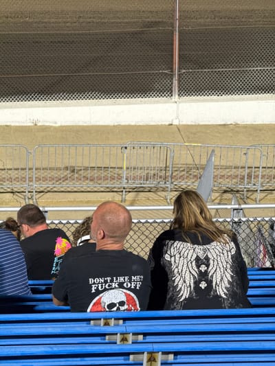 The image shows a rear view of several individuals sitting on blue bleachers, facing away from the camera. The setting appears to be an outdoor event, possibly a race track, with a fence and dirt track visible in the background.
In the foreground, on the right, a person with long blonde hair is wearing a black hooded sweatshirt with a large white winged design on the back. To their left, a bald-headed person in a black t-shirt has the text "DON'T LIKE ME? FUCK OFF" printed on the back, along with a graphic of a skull wearing a sombrero. Further to the left, a person wearing a blue and white striped shirt is visible. Another person with dark hair is seated between the person in the striped shirt and the bald-headed individual. The person to the left of the bald individual wears a black t-shirt with a pink and yellow palm tree graphic.
The bleachers are painted bright blue. In the background, a chain-link fence separates the seating area from what appears to be a dirt track or field. Another, higher fence with horizontal bars is behind that, and beyond that is a wall or barrier. The ground is covered with a tan-colored material, likely dirt or gravel. The overall atmosphere suggests a casual spectator event.