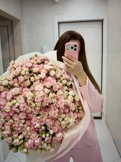 The image shows a person holding a large bouquet of pink and white roses. The person is holding a pink smartphone up to take a mirror selfie. The person is wearing a light pink jacket or sweater. The background appears to be an interior space, possibly a room or hallway, with a white door or panel in the reflection. The lighting suggests it is daytime. The overall mood is calm and celebratory, likely due to the flowers. The image is taken from a slightly high angle. There is no visible text in the image. The location context is Константиновка, Kyrgyzstan.