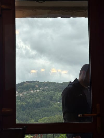 A person is partially visible, looking through a window at a landscape in Кирково, Bulgaria. The landscape shows a hillside covered in dense green trees and some buildings, under a cloudy sky. The window has a wooden frame, and the person is wearing a dark-colored shirt. Reflections are visible on the window surface.