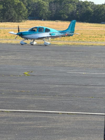 A small, light blue and white airplane is parked on an asphalt runway. The airplane has a single propeller at the front. The background consists of a grassy field with trees in the distance. The scene appears to be an airfield in the Town of East Hampton, United States of America.