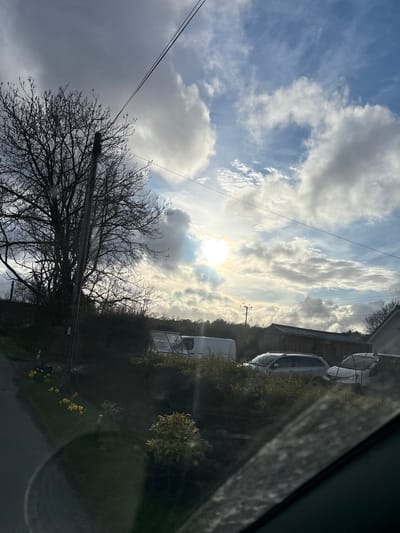 The image shows a view of the sky with scattered clouds and the sun shining through. A bare tree with many branches occupies the lower left portion. Utility poles and power lines are visible. On the right side, there are buildings, vehicles (possibly caravans), and vegetation, including some yellow flowers. The scene is likely outdoors. The image may have been taken from inside a vehicle, judging by the reflection or window edge visible on the right. The location is Shelve, United Kingdom.