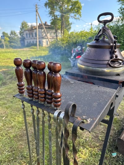The image, rotated vertically, captures an outdoor cooking scene in Луптюг, Russia, during what appears to be a bright, clear daytime. The primary subjects are a large, dark metallic pot, resembling a kazan or Dutch oven, resting on a black metal brazier or grill stand. Smoke billows gently from beneath the pot, indicating an active fire heating its contents. The pot has a sturdy handle and lid, with some unreadable embossed patterns or text on its side.
Arranged on the side of the brazier are several metal skewers, featuring distinctive twisted shafts and dark, polished wooden handles. These skewers are typically used for grilling meat, such as shashlik.
The background reveals a rural or semi-rural setting, with vibrant green grass covering the ground and lush trees providing foliage. In the distant left, a traditional-style building, possibly an old house or estate, with visible windows and roof structures, is partially visible. A utility pole with power lines is also present. The clear sky with some clouds suggests pleasant weather.
No individuals are visible in the frame. The scene portrays an inviting outdoor cooking activity, likely preparing a meal such as a stew or soup in the pot, alongside grilled items using the skewers.