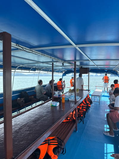 On a sunny day, several people are seated on a blue-decked boat under a blue canopy, likely in the waters near Khlong Khian, Thailand. The boat is equipped with brown benches lining the sides, and a long, wet wooden table runs down the center of the seating area. Numerous orange life vests are neatly stacked on shelves beneath the benches. Some passengers wear life vests, while others do not. The boat appears to be moving through calm, turquoise water, with distant, hazy islands visible on the horizon. The overall scene depicts a leisurely boat trip.