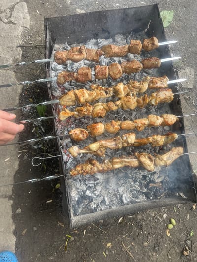 The image shows skewers of meat cooking over hot coals in a metal barbecue pit. The pit is filled with gray ash and glowing embers, with smoke rising from the coals. There are multiple skewers of meat, some appearing to be pork and others chicken, arranged in rows. A human hand is visible on the left side of the frame, seemingly adjusting or tending to the skewers. The barbecue pit is situated on dark soil, with some scattered leaves and small green plants. In the background, a concrete surface is visible, suggesting an outdoor setting. The overall scene depicts a barbecue in progress, likely in a residential or recreational area, possibly in Novyi Tahamlyk, Ukraine, given the context provided.