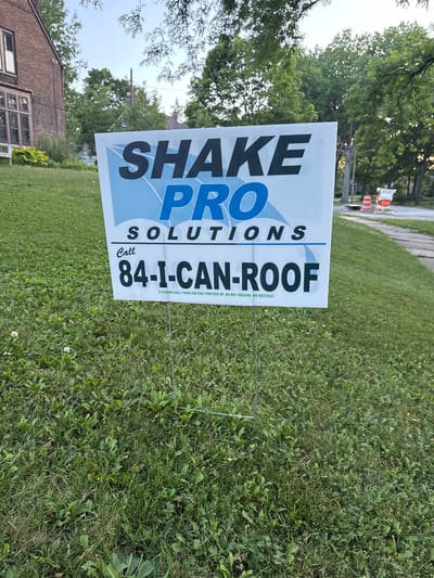A white yard sign with blue and black lettering stands in the foreground of a residential yard. The sign displays the words "SHAKE PRO SOLUTIONS" in large, bold lettering, with a phone number "84-I-CAN-ROOF" underneath. The sign is supported by two thin metal stakes. The yard is covered in green grass with scattered clover and weeds. In the background, a brick house with multi-paned windows is visible, along with several mature trees and a sidewalk. Further in the background, traffic cones and construction barriers are present. The sky above is a pale blue with a few faint clouds. The location is Shaker Heights, Ohio, United States of America.