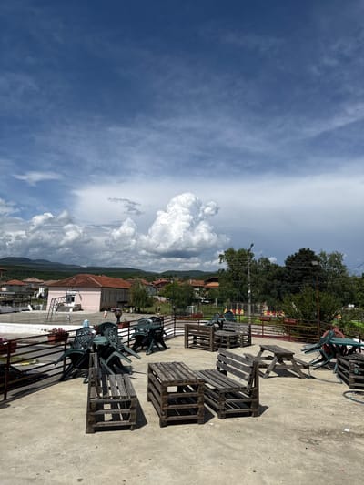 This image captures an outdoor scene in Blatska, Bulgaria, likely a terrace or a large paved area, under a bright, partly cloudy sky. The foreground features a concrete or paved ground surface. Arranged across this surface are numerous wooden pallets, some stacked to form benches and tables, suggesting a makeshift seating area. Several green plastic chairs, some stacked or overturned, are also present among the pallets. A metal railing or fence runs along the edge of this paved space, visible on the right side of the image (when rotated correctly). A blue hose or pipe is coiled on the ground, and scattered pieces of light-colored litter or paper are visible.
In the middle ground, beyond the railing, residential or commercial buildings with red-tiled roofs and light-colored facades (including a prominent pink building) are nestled amidst lush green trees. A street light or utility pole with overhead wires rises above the trees. Further in the distance, a dark green soccer goal net can be faintly discerned. The background reveals rolling mountains under a dynamic sky filled with large, fluffy white cumulus clouds, particularly dense on the left side, contrasting with vast expanses of blue sky and some fainter wispy clouds.
The scene depicts a calm and empty environment, with no people visible, suggesting it might be an off-peak time or a closed establishment. The bright sunlight and distinct shadows indicate it is daytime, likely late morning or early afternoon. No legible text is visible in the image.