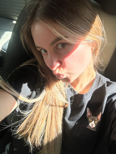 A woman with blonde hair is taking a selfie in a car, possibly as a passenger. She is wearing a black t-shirt with a cat design on the pocket. Sunlight is casting shadows on her face and hair. The background appears to be the interior of a car, with seats visible. There are no discernible details about the city of L'Essard, France.