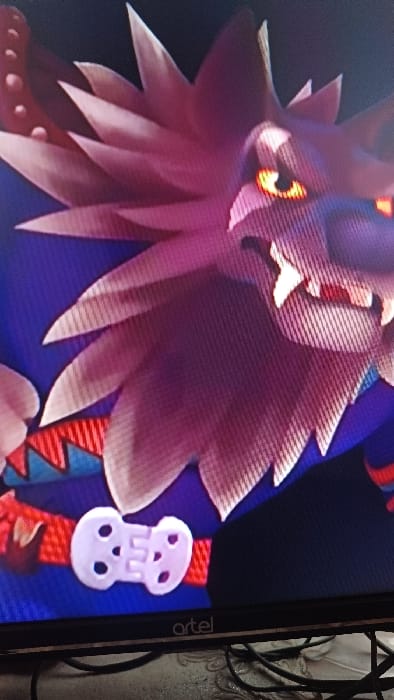 The photograph captures a close-up of a digital display, identified by the "artel" logo at the bottom edge, exhibiting an animated character. The character is a stylized, anthropomorphic creature resembling a wolf or lion, rendered in shades of purple and pink with spiky fur or a mane. Its prominent features include glowing yellow-orange eyes and visible fangs. The character is depicted wearing a red belt with a white, bone-shaped buckle or ornamental piece, featuring two circular cutouts on each side of a central "E"-like shape. The immediate environment beneath the display shows tangled black cables resting on a light-colored, patterned surface. The image was reportedly captured at a location within the city of Kasansay, Uzbekistan.