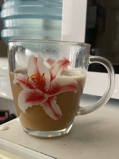 A close-up, eye-level shot captures a clear glass mug filled with a light brown beverage, likely coffee with milk, sitting on a white surface. The mug is adorned with a decal of a pink and white lily flower with orange stamens. Condensation droplets are visible on the exterior of the mug. In the background, a large, blue, translucent water cooler jug is out of focus. To the right of the mug, a dark, indistinct object is visible. The overall setting appears to be indoors, possibly a kitchen or office, on the territory of Salsk, Russia.