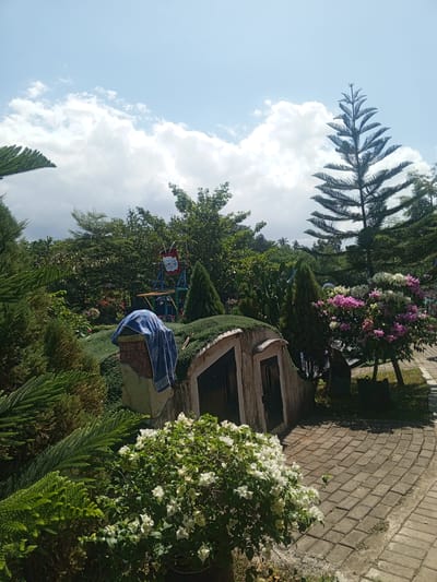 This is a wide shot of a garden with a blue sky and white clouds. In the foreground, there is a small, arched building covered in green foliage. To the left of the building, a blue towel is draped over a brick structure. There are various plants and trees surrounding the building, including cypress trees, flowering bushes with pink and white flowers, and a tall pine tree. In the background, a red and white sign is visible on a metal structure. The ground is paved with brick tiles. This image appears to be taken in Darsono, Indonesia.