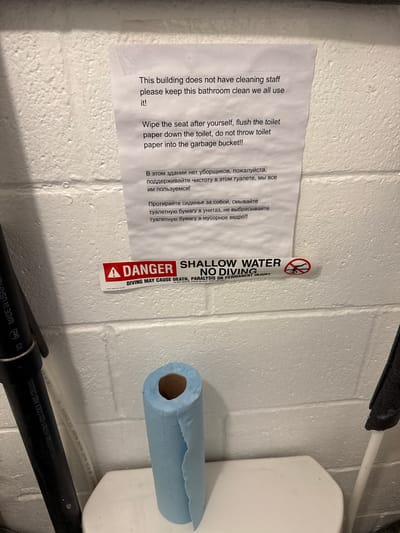 The image depicts a bathroom scene with a white cinder block wall. A white piece of paper with instructions is taped to the wall. Below the paper, a red and white "DANGER" sticker with a "NO DIVING" symbol is also affixed to the wall. A blue paper towel roll sits on the toilet seat. To the left of the toilet, a black pipe with white lettering is visible, and to the right, a black padded object is partially visible. The environment appears to be a public or communal restroom. The provided context indicates this scene is located in the city of Richmond Hill, Canada.