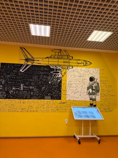 The image captures an interior space in what appears to be a corridor or waiting area, possibly within an educational institution or a science-themed environment. The dominant visual feature is a large, yellow wall adorned with hand-drawn illustrations in black ink, depicting a rocket, an astronaut, and numerous mathematical and scientific formulas. This mural gives the impression of a chalkboard filled with equations and diagrams.
On the right side of the image, a digital display screen on a white mobile stand is situated against the yellow wall, which is also next to an orange wall. The screen displays a digital interface with a grid-like arrangement of boxes and text.
The wall to the left is characterized by a grid-like pattern, suggestive of a ceiling or architectural element with embedded lighting fixtures that are turned on, illuminating the space. The room lighting casts brightness onto the foreground and mural.
Based on information regarding a space with similar features, it is plausible that this location is within the city of Murmansk, Russia. This information is derived from external sources and not inherently deducible from the image's content.