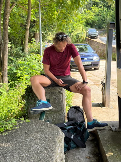A man is sitting outdoors, leaning against a stone structure, with his legs extended. He is wearing a maroon t-shirt, dark shorts, and blue athletic shoes. The man is holding a smartphone in his right hand and appears to be looking at it. His left hand is resting on his left knee. He is also wearing sunglasses pushed up on his head. A black backpack is on the ground behind him, partially obscured by a dark green bag.
In the background, there are trees and lush green foliage. Two cars are visible on a road behind the man: a dark-colored SUV and a blue Volkswagen hatchback. The license plate of the Volkswagen is visible and reads "T27-E-006". The scene appears to be set during the daytime, with sunlight casting shadows. The overall mood seems relaxed, as the man is taking a break. The location cues, such as the cars and the surname "Jajce" in the provided context, suggest the setting is Jajce, Bosnia and Herzegovina.