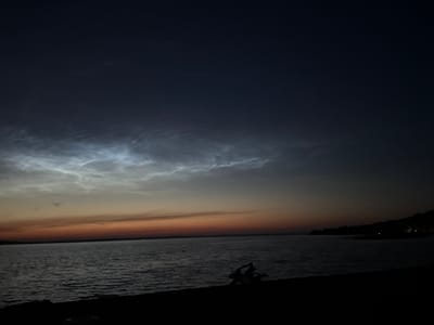 The image depicts a twilight scene over a body of water, likely a lake or river, with a dark shoreline in the foreground. The sky displays a gradient of colors, transitioning from deep blue at the top to a muted orange and pink near the horizon. Wispy, illuminated clouds, possibly noctilucent clouds, are visible in the upper portion of the sky. The water is dark and reflects the ambient light. On the shoreline in the lower right quadrant, there is a dark silhouette of what appears to be a person or group of people, possibly engaged in an activity, and another dark object, possibly a piece of driftwood or debris. In the distance, along the horizon, there are faint lights indicating a settlement or structures. The overall atmosphere is serene and captures the transition from day to night. The location is identified as Володарский ауылдық округі, Kazakhstan.